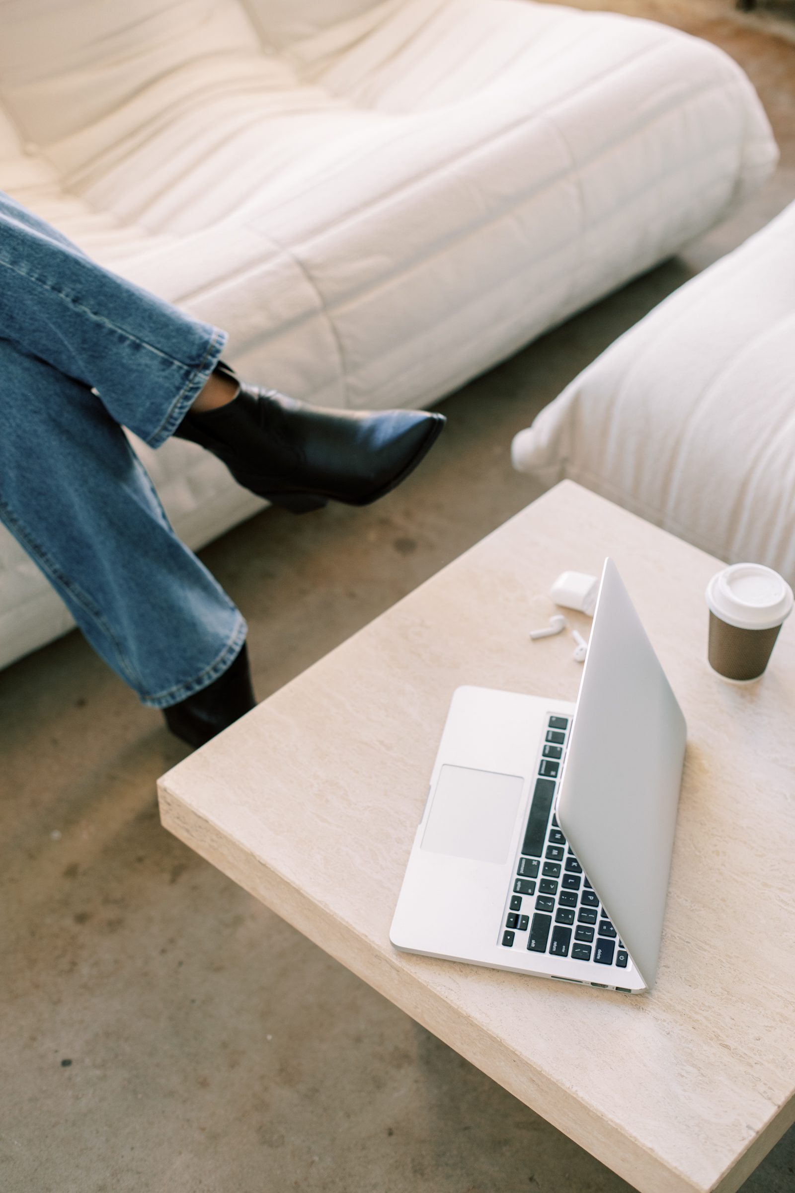 Woman in black boots and jeans sitting on a white table. In front of her is a coffee table with an open laptop and cup of coffee.